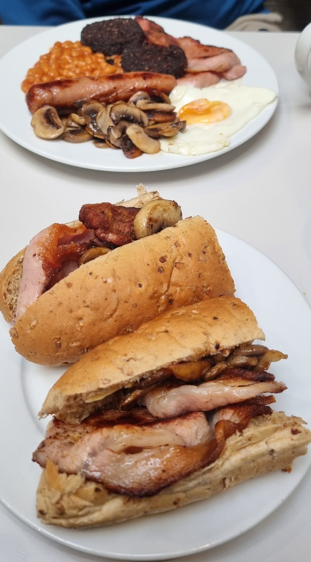 Two bacon and sausage rolls with a cooked breakfast plate behind